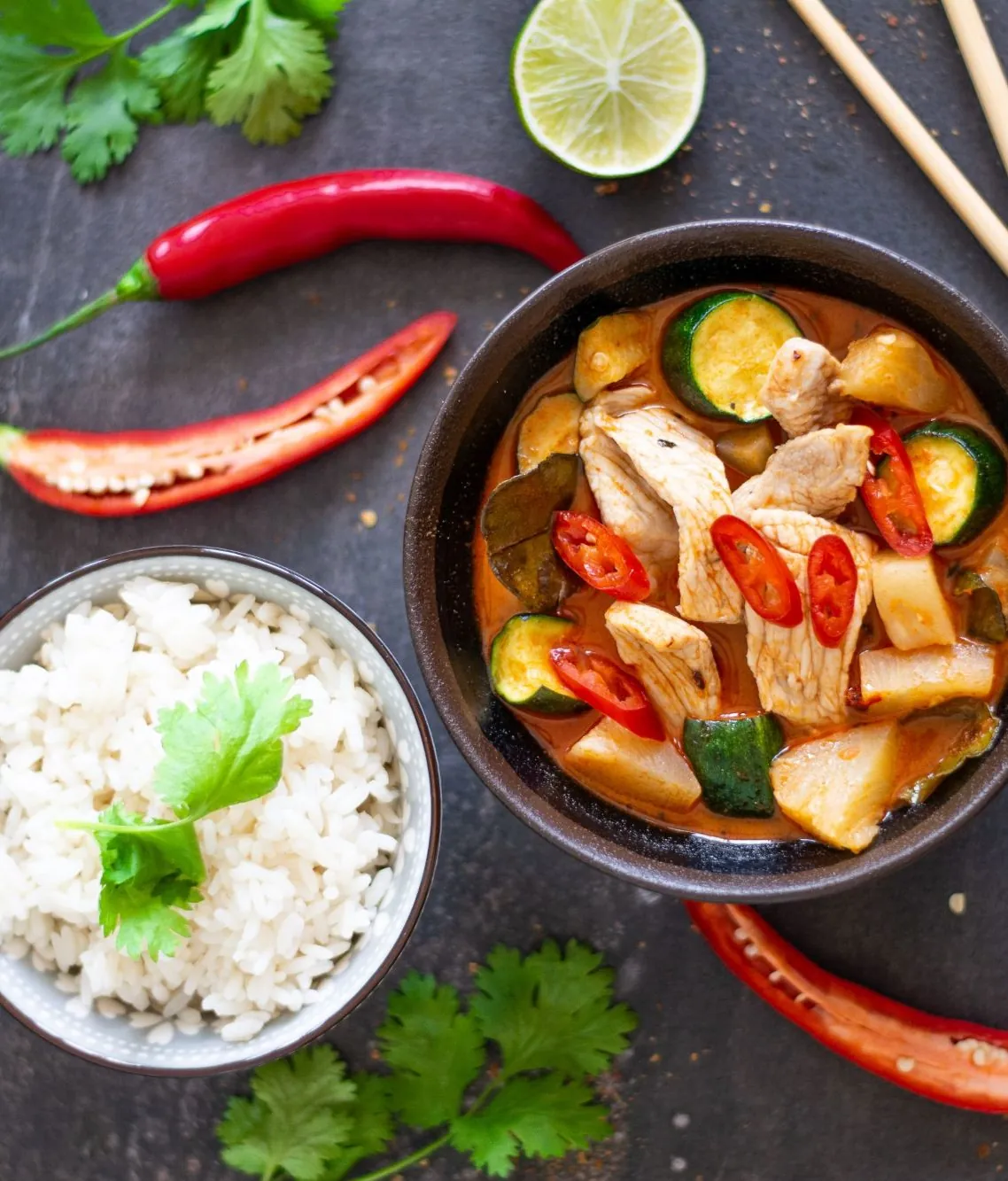 A bowl of red curry with chicken and courgette, with a bowl of rice and a lime beside it