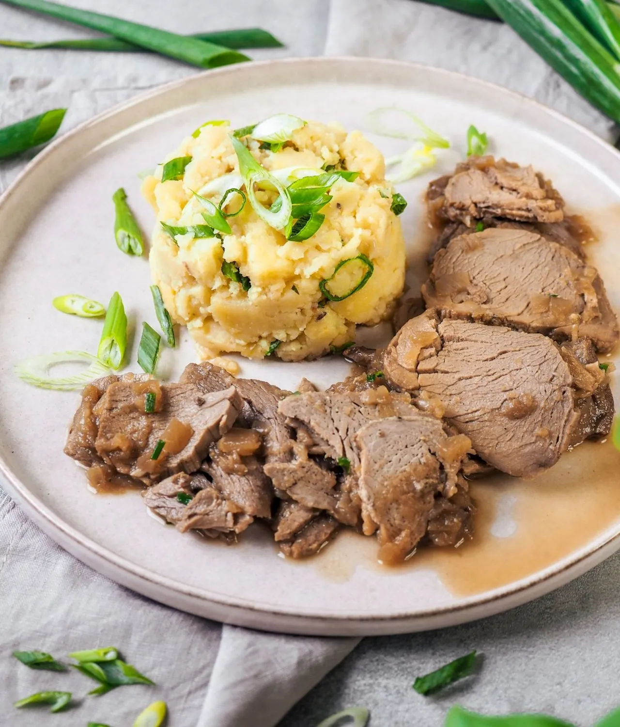 A plate of sliced pork tenderloin with crushed potatoes and spring onion